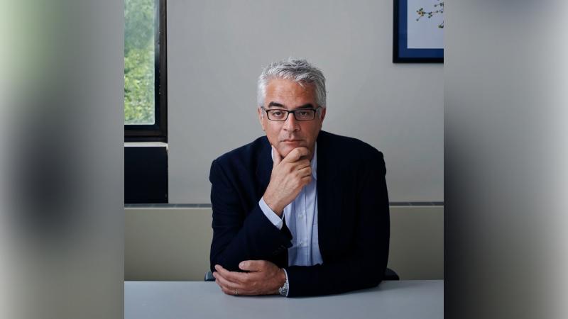 A middle-aged man with glasses and gray hair sits at a table, thoughtfully resting his chin on his hand, with a window and framed artwork in the background.