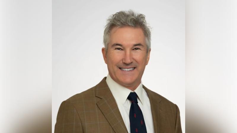 A smiling man with gray hair in a tan plaid blazer, white shirt, and navy tie with red designs, set against a light gray background.