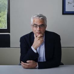 A man with glasses and gray hair sits at a table, thoughtfully resting his chin on his hand, with a window and framed artwork in the background.